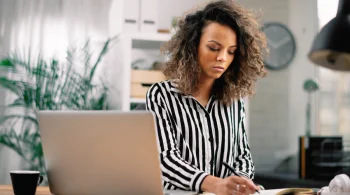 Woman writing notes beside a laptop at home