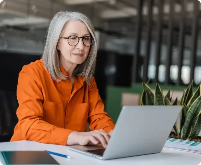 Older White woman with grey hair wearing glasses and a long sleeved orange shirt working on her LegalZoom corporate dissolution on her laptop.