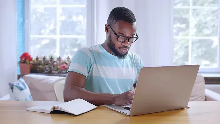 Young man at a bright workstation with an open notebook on desk, working on his laptop and reading about how to get a business license in Illinois.