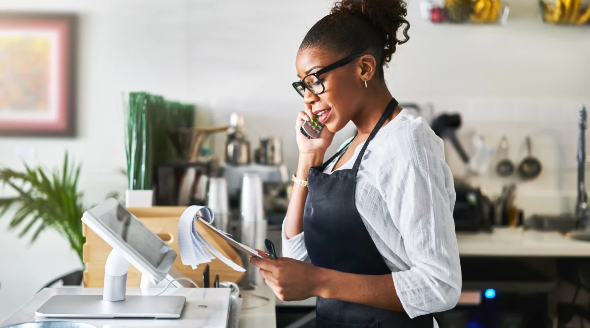 A woman stands at the counter in her bakery and reviews an order sheet while she speaks on the phone.