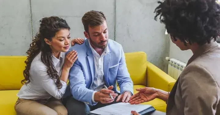 A couple sits on a yellow couch, signing trust documents with an attorney.
