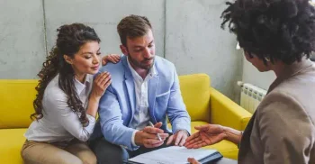 A couple sits on a yellow couch, signing trust documents with an attorney.