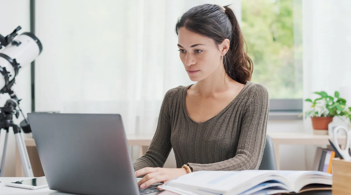 Woman working on a laptop with a telescope nearby