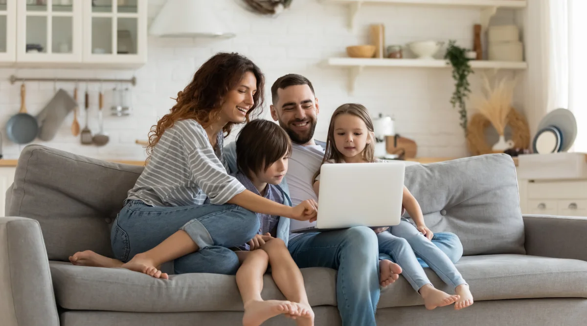 Family of four in a playful mood, sitting on the couch, all smiles, with the father holding the laptop.