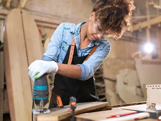 Woman in a denim shirt and a black apron using a power tool to cut a piece of wood for a sign for her business.