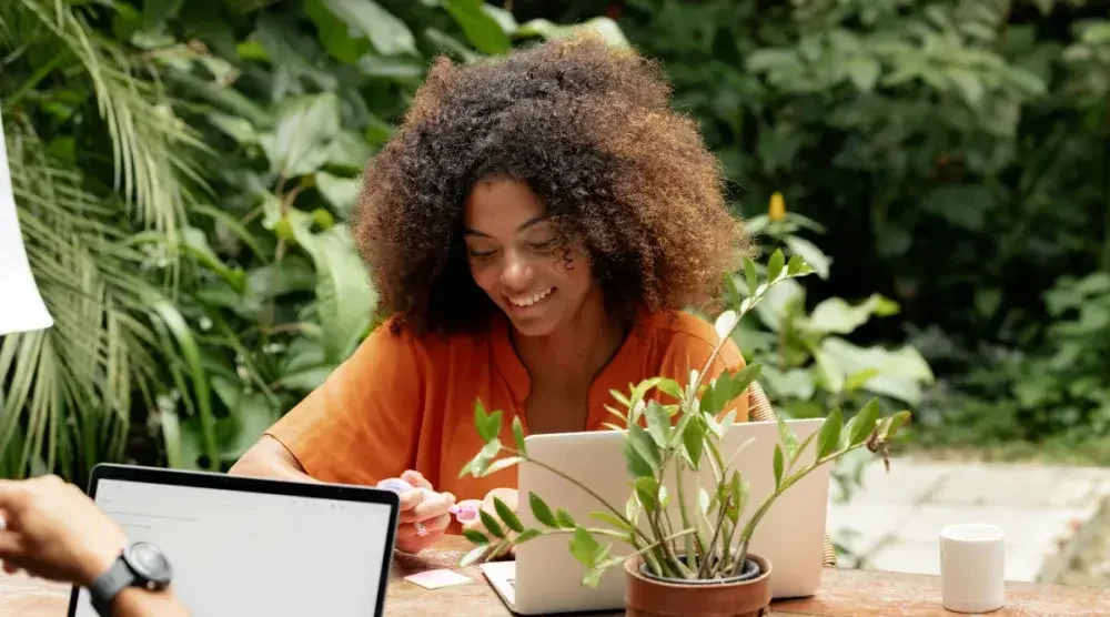 A woman sits at a table in her garden working on her laptop.