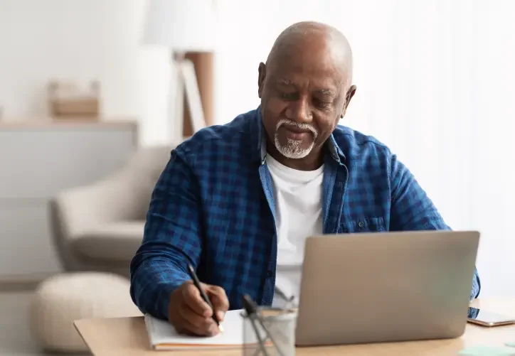 An older bald man with a white goatee and checkered shirt writes notes to himself while on his laptop.