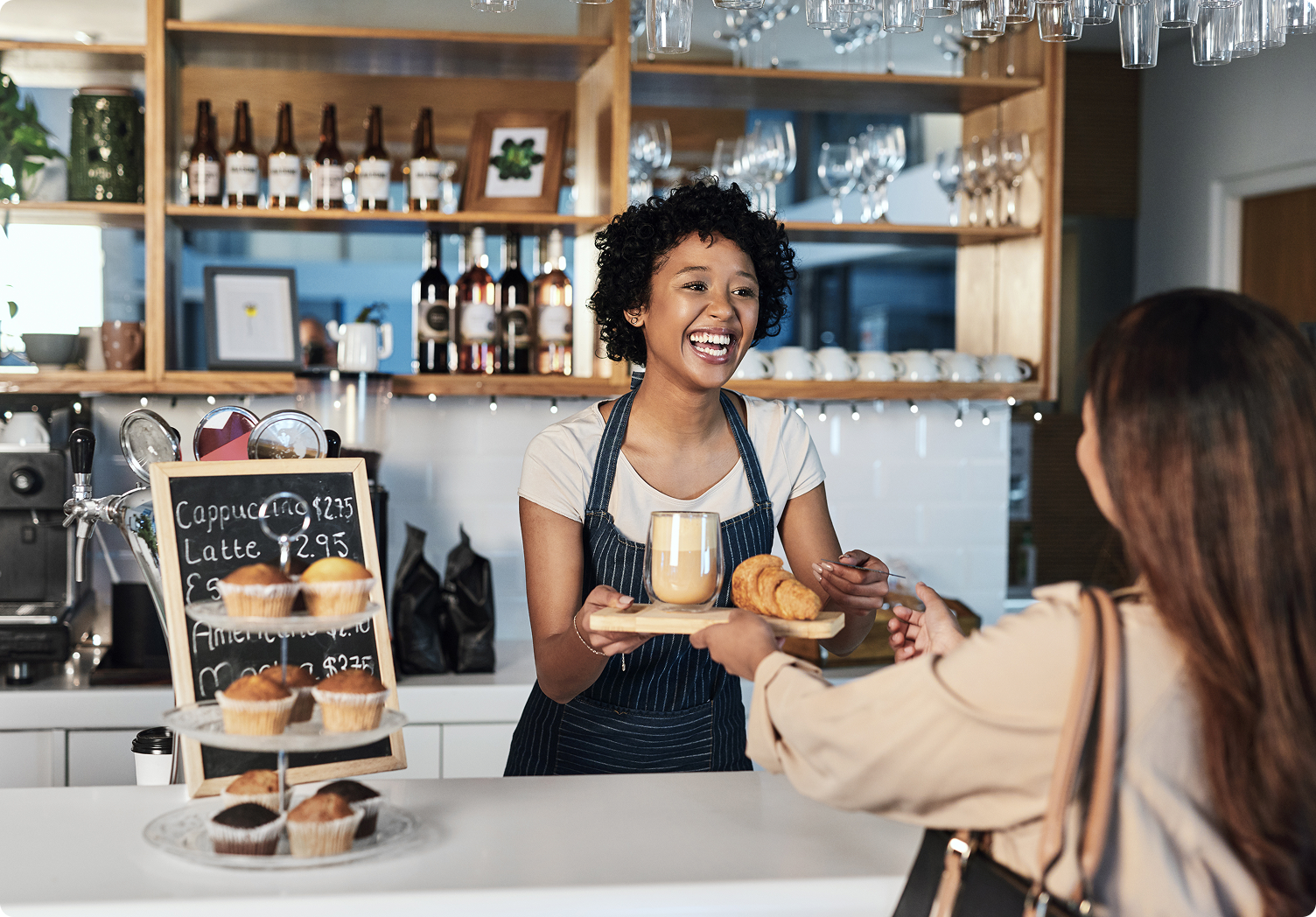 Black woman with curly hair wearing a white t-shirt and a blue and white pinstriped apron working in a café and  smiling because she got her business license from LegalZoom as she hands a woman with brown hair wearing a tan shirt her order.