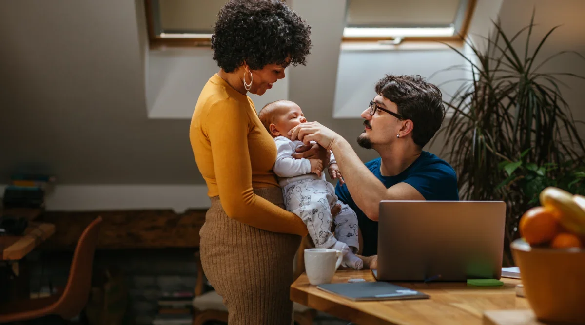 A young family stands in their living room. The mother holds their sleeping baby while the father touches the baby's face.