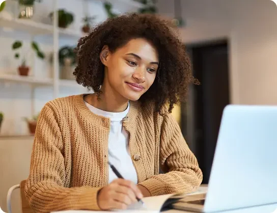 A young woman with curly hair, wearing a beige sweater, sits at a table writing in a notebook while looking at her laptop