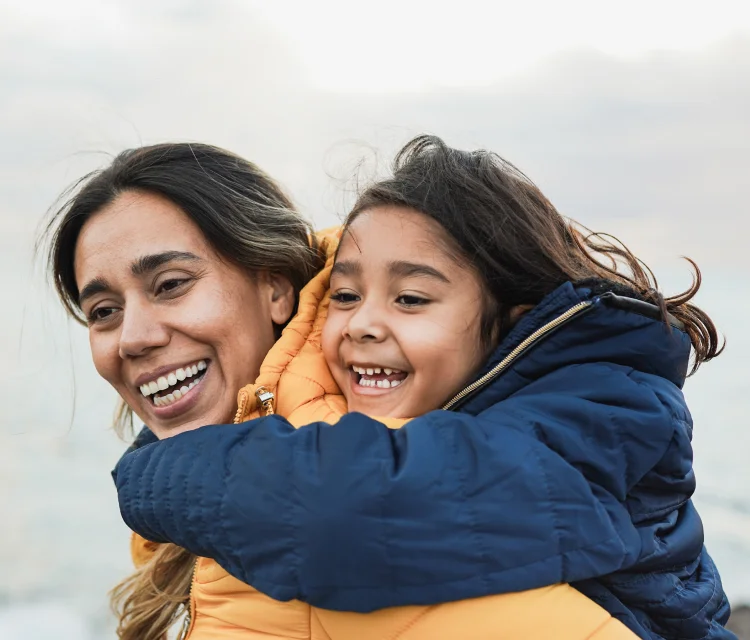 A woman with dark hair and a big smile and carries her daughter on her back. They are laughing.