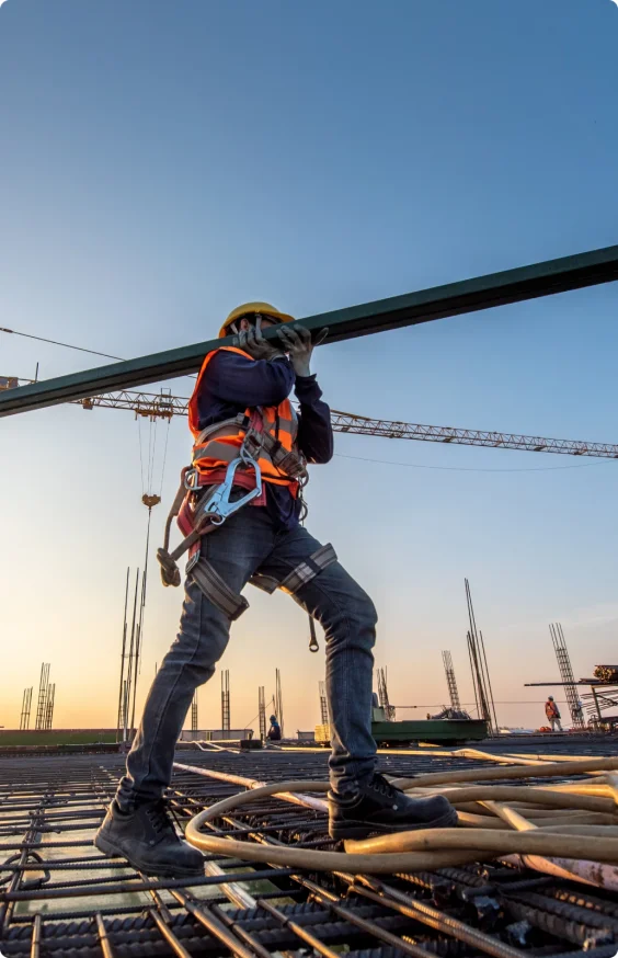 A construction worker in a high visibility vest carries a metal beam on his shoulder, through an jobsite.