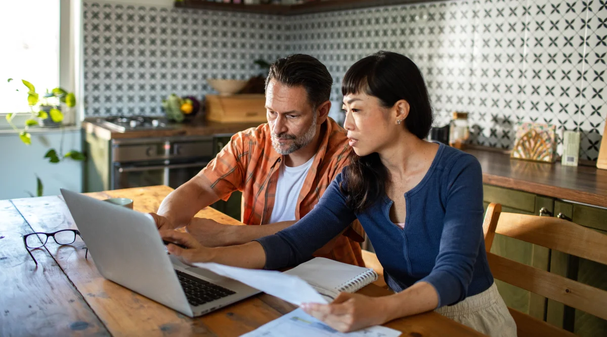 A man and woman sit at a table and look at a laptop screen.