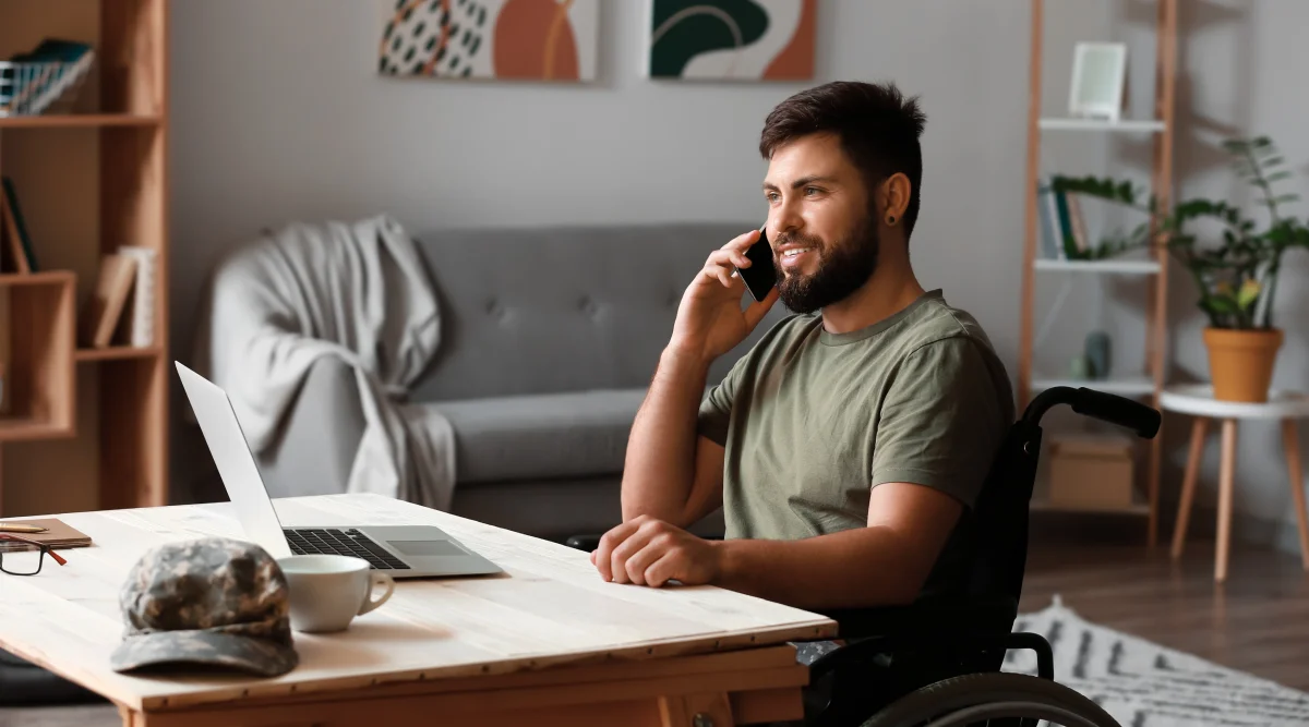 A man in an olive drab shirt sits in a wheelchair at a desk in his sunny apartment, smiling as he talks on the phone.