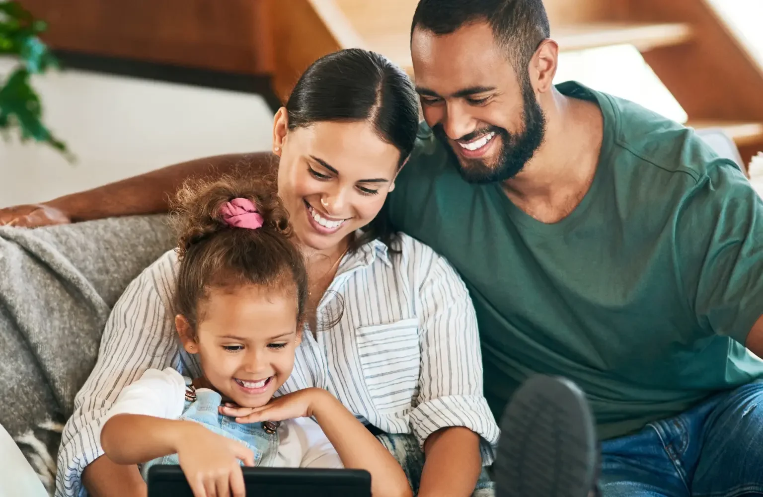 A family smiling and looking at a tablet. Mom, a woman with dark hair in a ponytail wearing a striped shirt, has the daughter in her lap. The dad is wearing a dark green shirt and has his arm around the mom's shoulder.