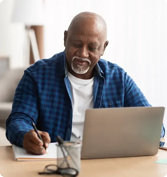 Bald man with a blue checked shirt writes a note while at his laptop