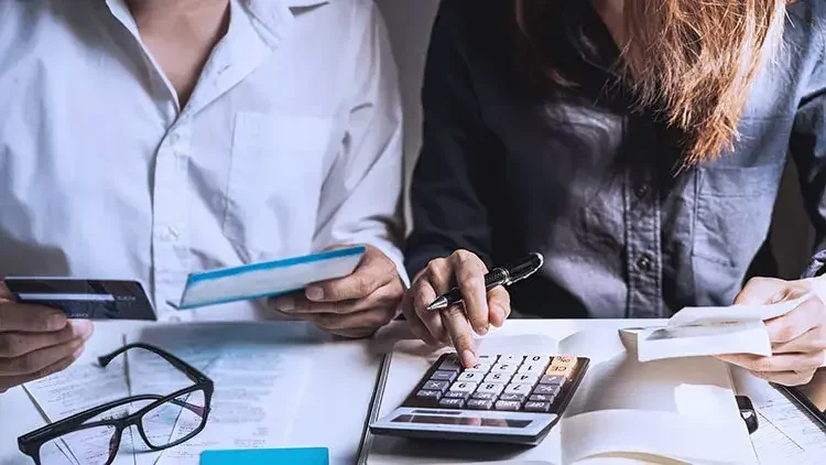 Two people sit at a desk, we see just their torsos and hands using a calculator and holding cards and a pen.