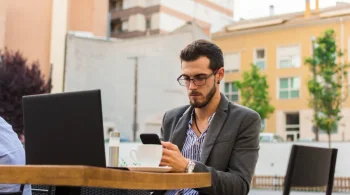 Man checking his phone at an outdoor cafe table with a laptop