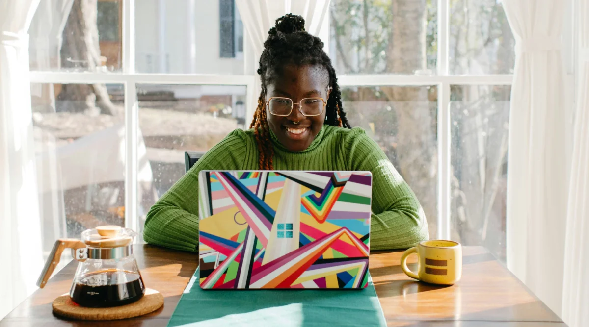 A woman in a green sweater smiles at her colorful laptop while enjoying coffee at a table