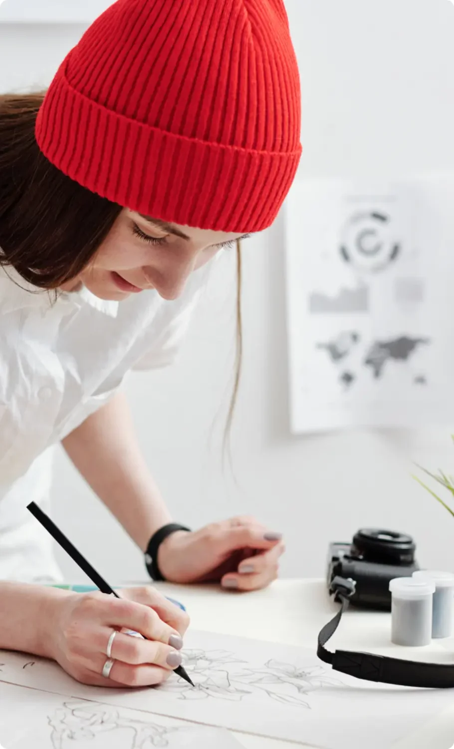 Woman in a shirt and red beanie writes with a pencil on her desk.