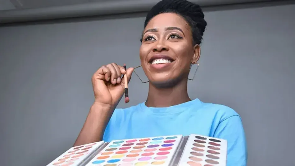 A makeup artist who owns an LLC in NY holds a palette in one hand and a makeup brush in the other hand.