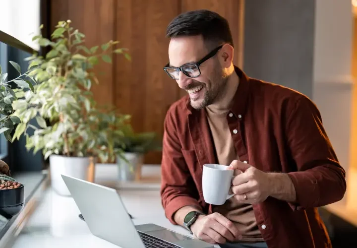 A man with glasses and a red button up shirt drinks coffee and smiles at his laptop.