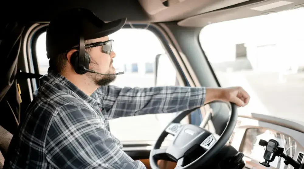 Profile of a man in a hat, sunglasses, and headset driving a truck.