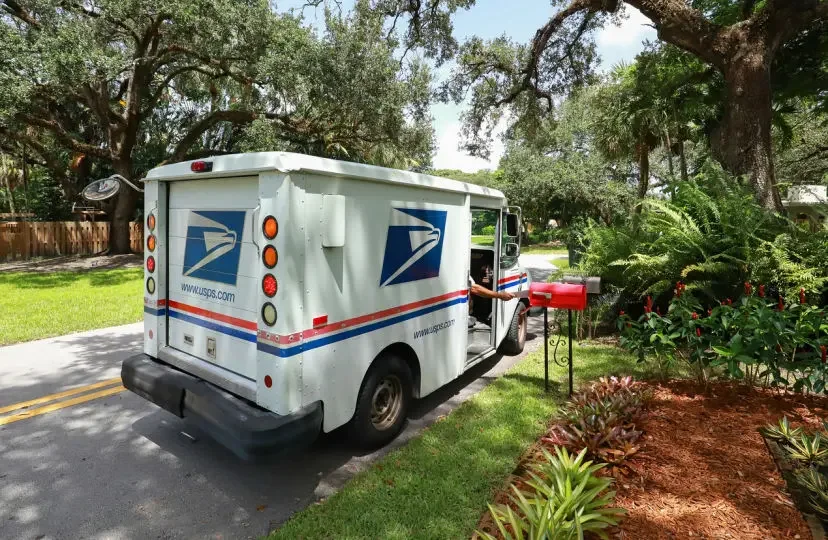 A person in a U.S. Postal Service truck reaches into a mailbox on a rural or suburban road. The USPS delivers two types of packages on Sundays.