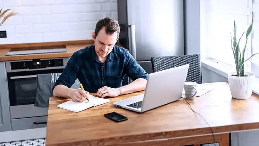 An image of a man writing in a notebook at the kitchen table.