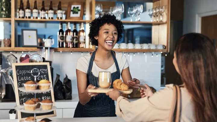 Black woman with curly hair wearing a white t-shirt and a blue and white pinstriped apron working in a café and  smiling because she got her business license from LegalZoom as she hands a woman with brown hair wearing a tan shirt her order.