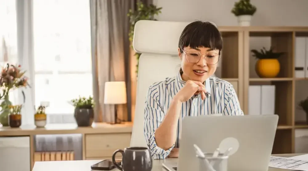 A woman seated at a desk rests her chin on her hand while she looks up BOIR on her laptop. 