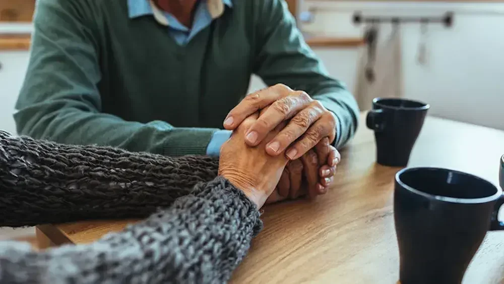 An elderly couple holds hands at a kitchen table. Executing a trust, including appointing a trustee and choosing beneficiaries, is crucial to ensure the trust serves its intended purpose and protects your legacy and your voice.