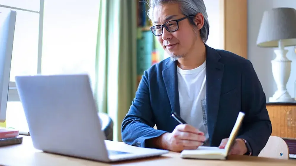 A grey-haired man in glasses sits in front of his laptop while writing in a notebook.