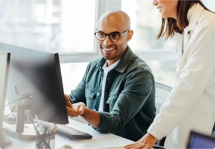 man wearing glasses smiles at his desktop, and speaks to his coworker.