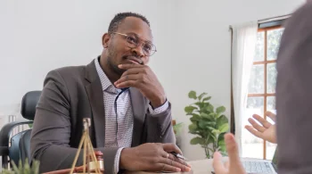 A man in a suit thoughtfully listens during a conversation in a bright office setting