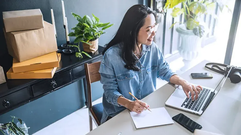 A woman sits at her office desk, smiling as she types on a laptop and takes notes. 