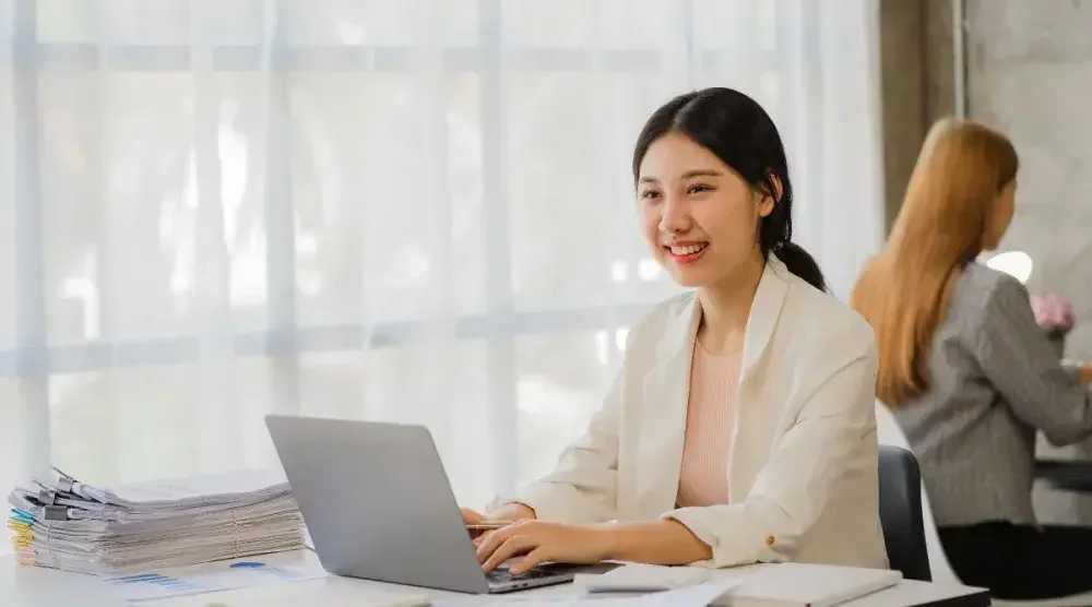 A woman sits at her desk in a bright, open-concept office and types on a laptop.