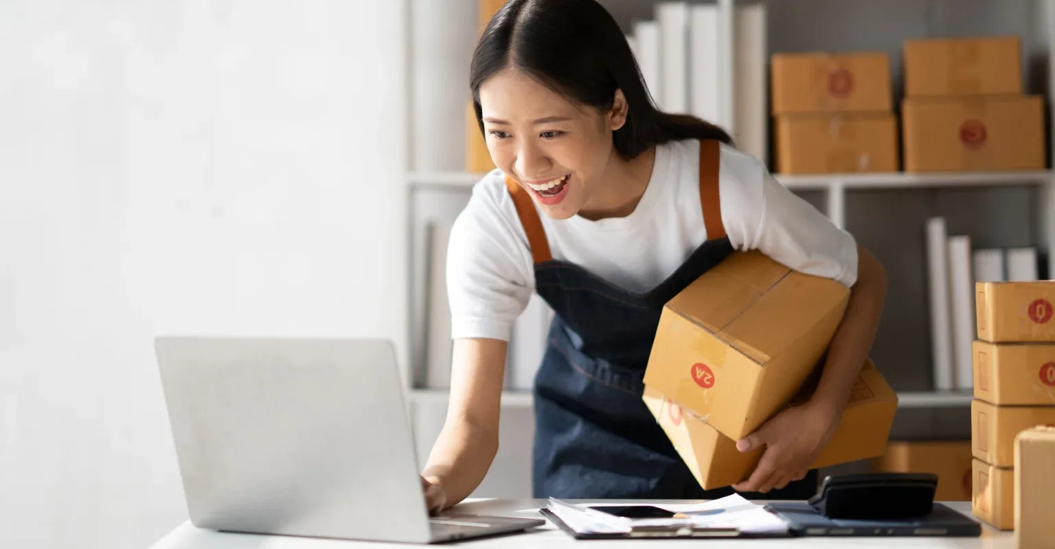 Asian woman wearing a white t-shirt and a denim apron is smiling as she ships packages, because she formed her LLC with LegalZoom