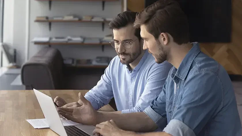 2 men wearing blue shirts looking at a laptop and talking.