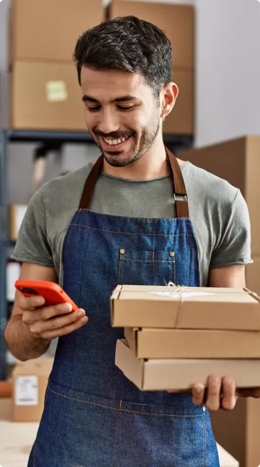 Smiling young man wearing a gray shirt and denim apron standing in a warehouse with boxes in the background looking at his cell phone in one hand and holding packages in the other.