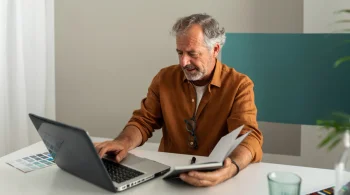 A man with gray hair studies documents while working on a laptop in a stylish office space