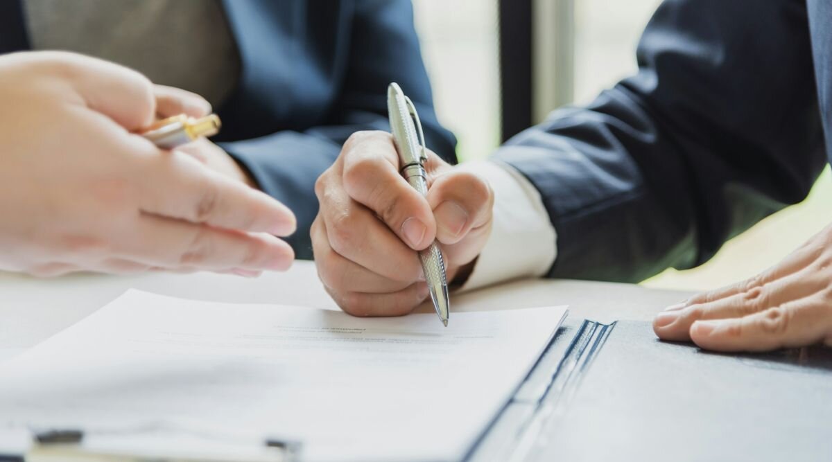  Three people in business suits sit at a large glass table, discussing and signing a contract.