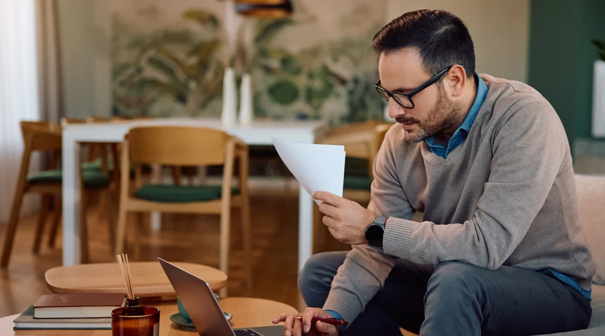 Man reading paper, working on a laptop in a cozy room