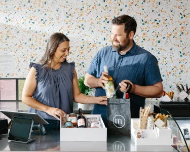 Brunette white woman wearing a blue ruffled tank top helping a brunette White man with a beard wearing a blue button up shirt packing a wine shipment after getting their business license through LegalZoom.