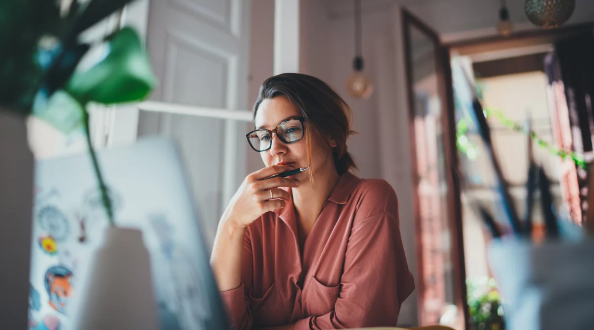 A woman ponders thoughtfully, holding a pen, while looking at her laptop in a bright room