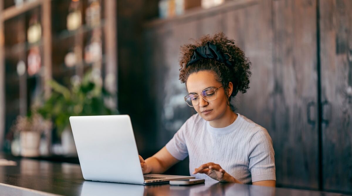 A woman sits in an open space cafe and checks something on her phone while using a laptop.