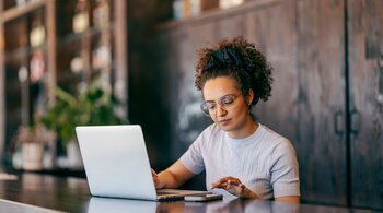A woman sits in a cafe and uses a laptop and a phone.