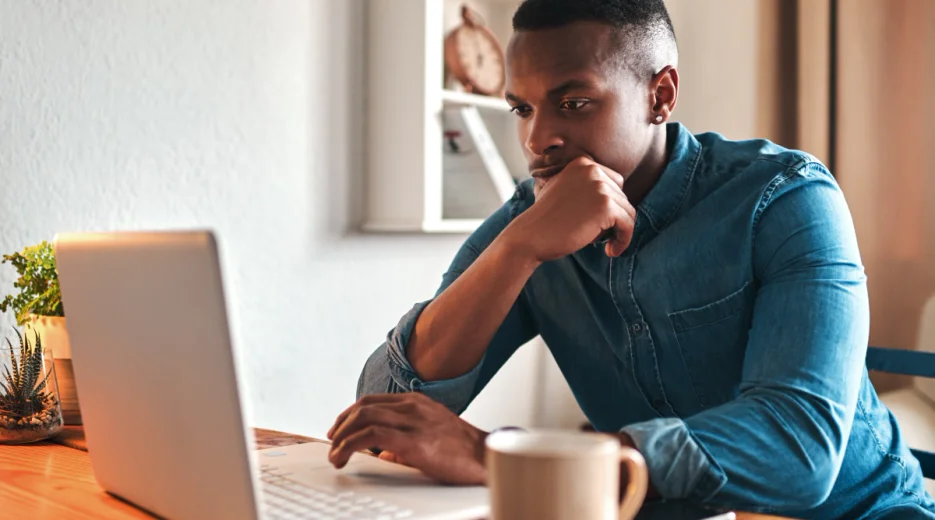 A man is sitting at a table with a coffee mug, reading about a spendthrift trust on his laptop.