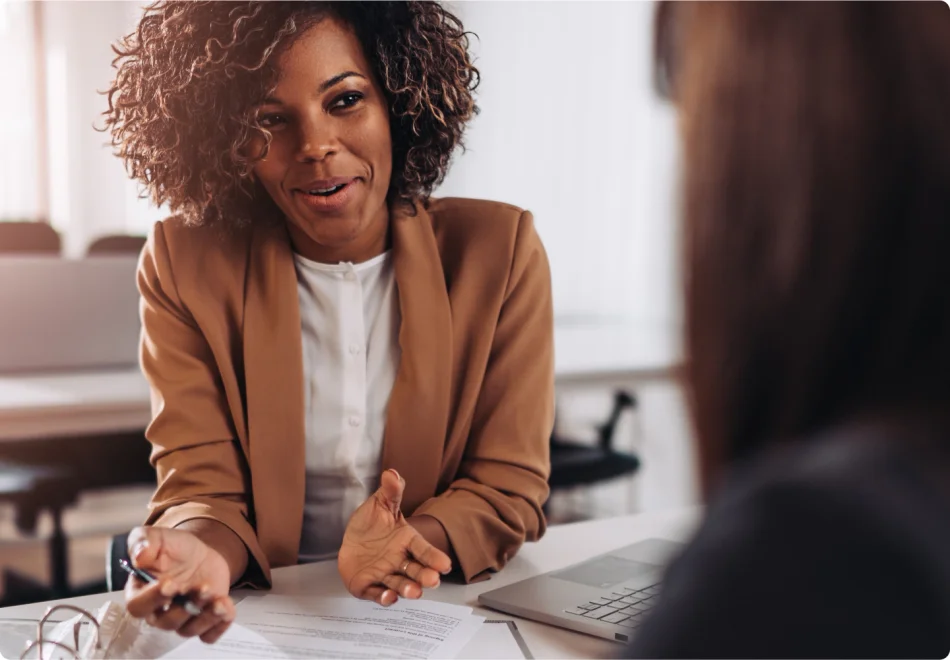 A black woman wearing a brown business suit has a discussion over paperwork with a client.