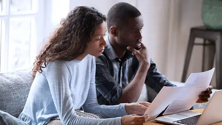 A man and a woman sit next to each other on a couch, glancing at documents with a laptop on the desk in front of them. 